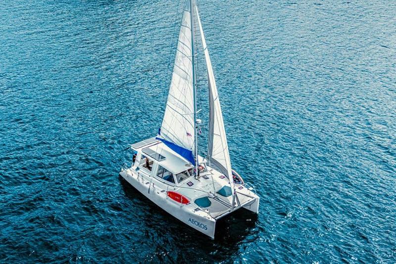 An aerial view of a catamaran named AEOLIS sailing on the sea with its sail raised, cutting through 
