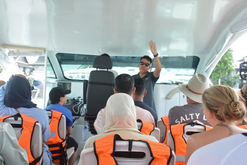 A tour guide giving a briefing to passengers wearing orange life jackets inside a speed boat, with c