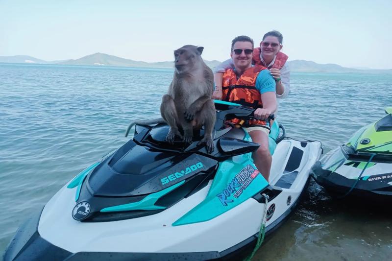 A monkey sitting on a jet ski with two people in life jackets on a beach in Phuket, Thailand.